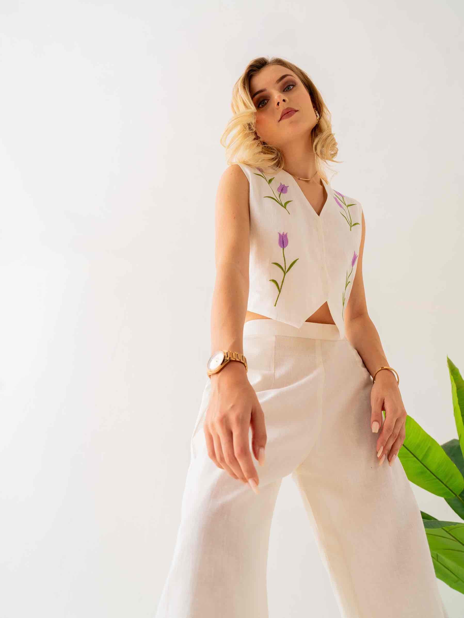 Woman wearing a white sleeveless top with floral embroidery and beige pants against a white background.